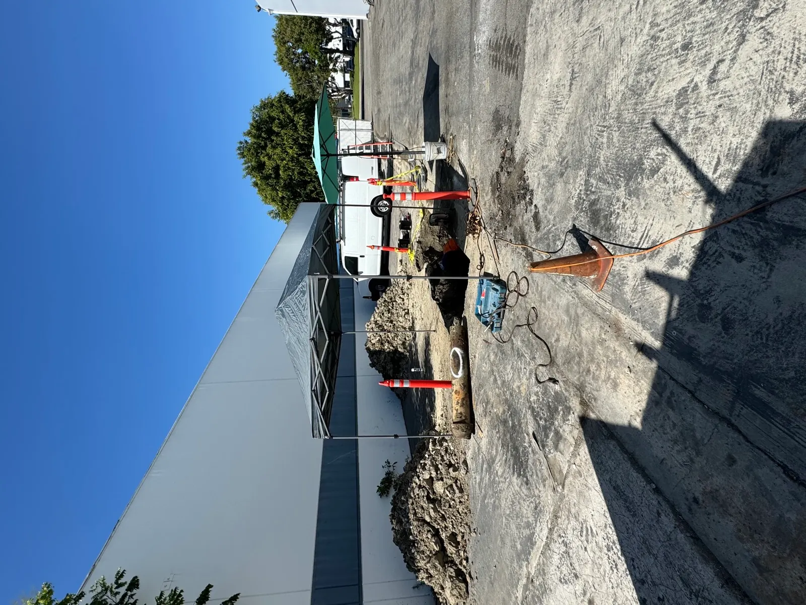 Excavated sewer repair under a canopy in a commercial parking lot with dirt piles and traffic cones