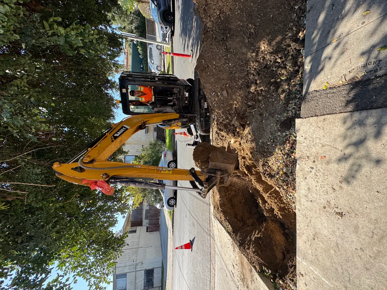 SANY mini excavator digging a sewer lateral trench at the curb line with a Trifecta operator